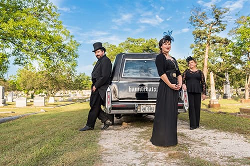 Three people dressed in all black formal attire standing around one hearse in a grave yard